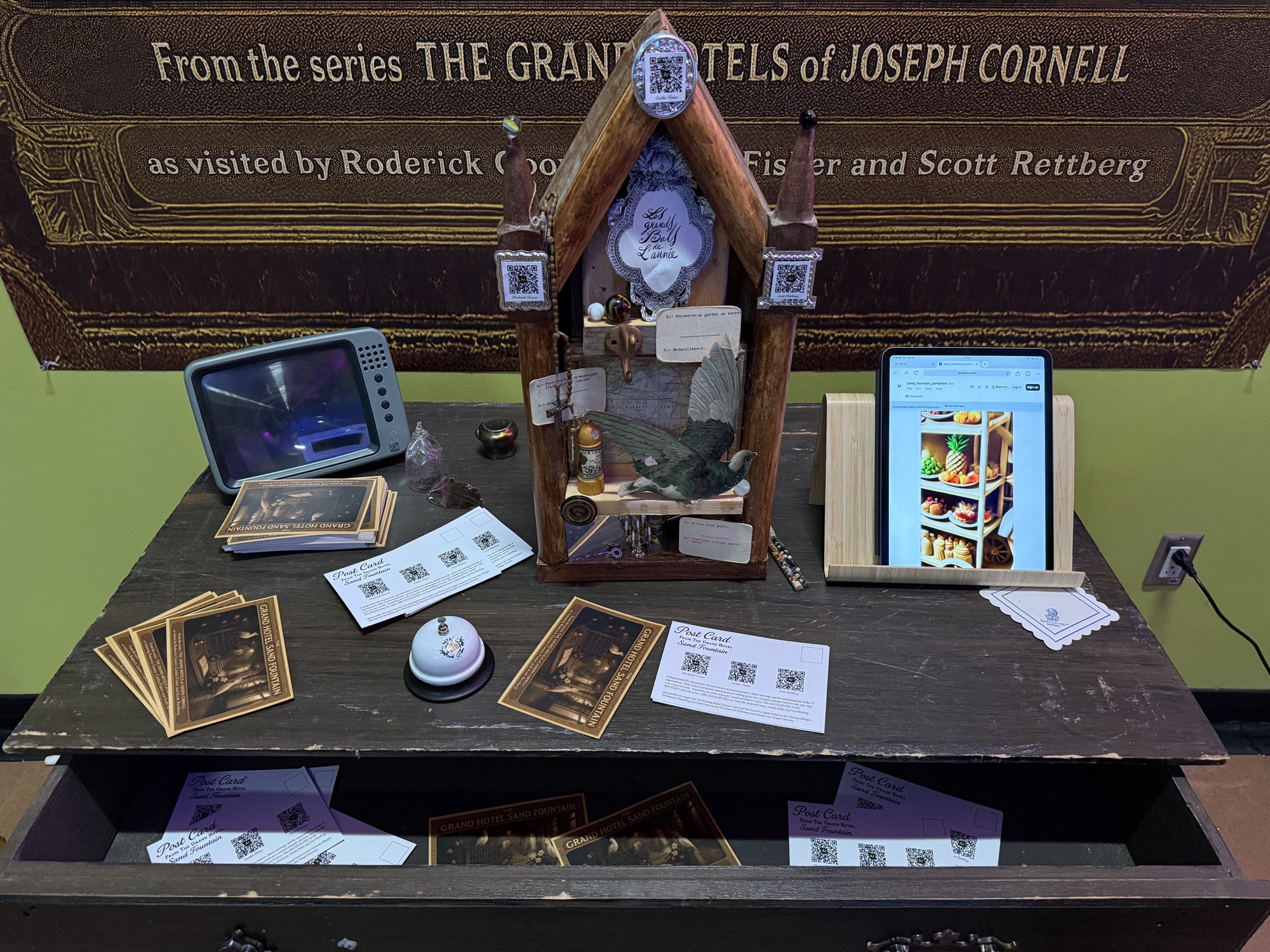 A close view of The Grand Hotel Sand Fountain installation with collage, tablet, postcards, bell, and video display at ELO 2025.